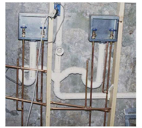 Rough-in of an indoor plumbing system: copper water lines and white PVC drain pipes mounted on a concrete wall with two blue valve boxes at the top.