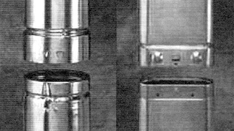 Row of tall stainless steel containers or dispensers mounted on a dark wall in a utilitarian kitchen setting.