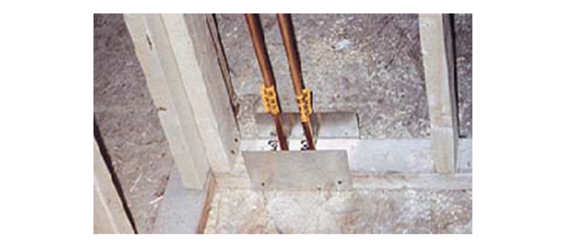 Two copper water pipes with yellow tags pass through a metal plate in a concrete opening inside a wooden-framed wall under construction.
