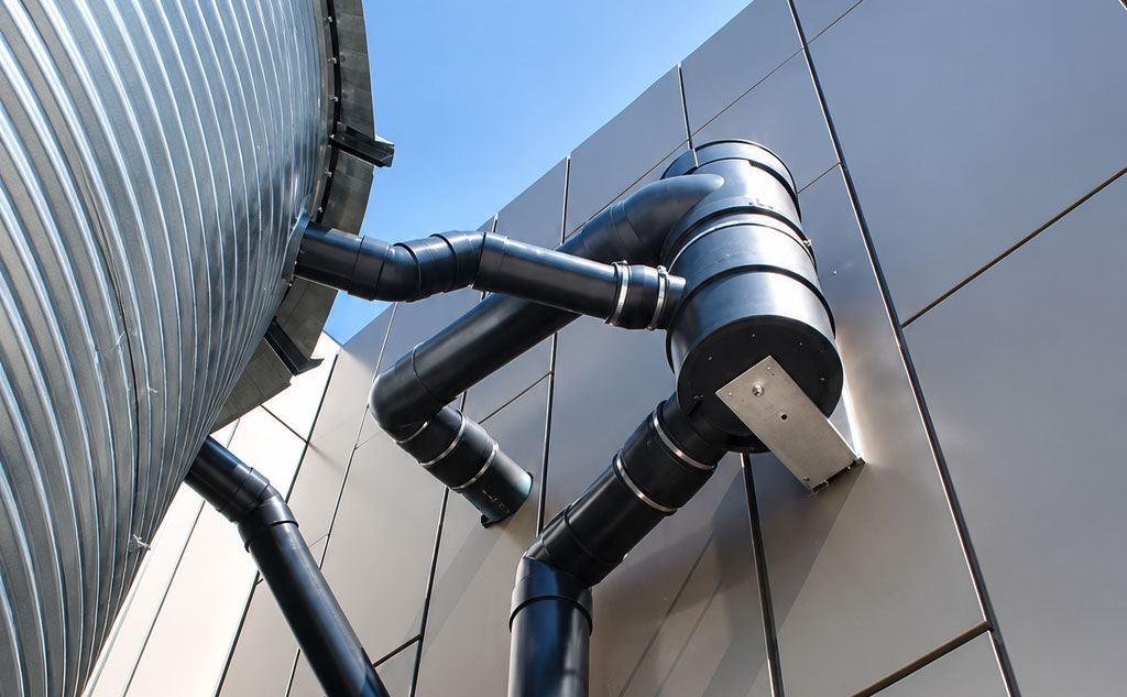 Industrial pipes and vents mounted on a silver building against a blue sky.