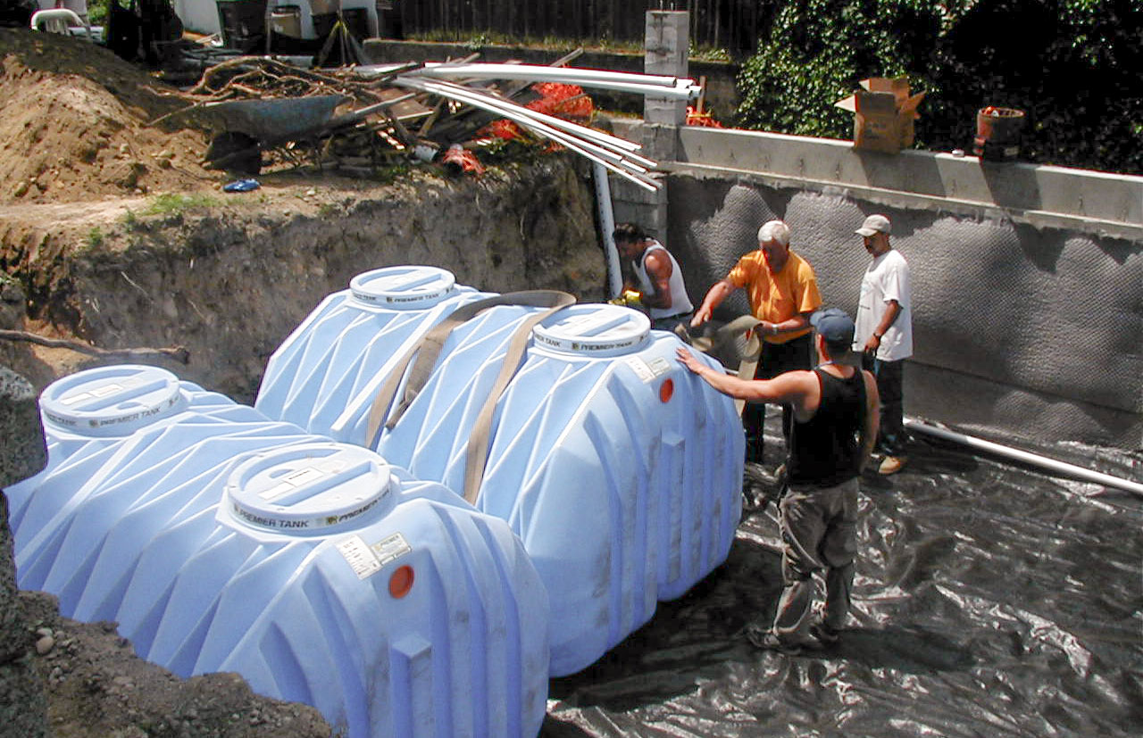 Construction workers install large blue water tanks in a trench lined with plastic sheeting