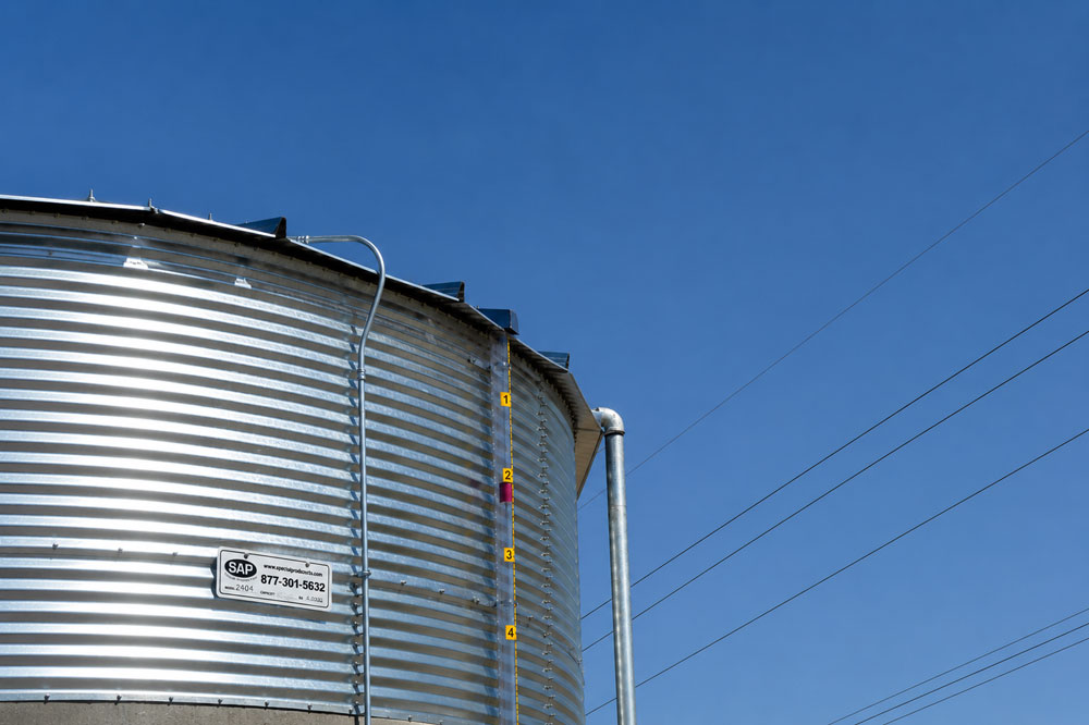 Silver corrugated grain silo with a vertical yellow measurement scale against a clear blue sky; power lines in the distance.