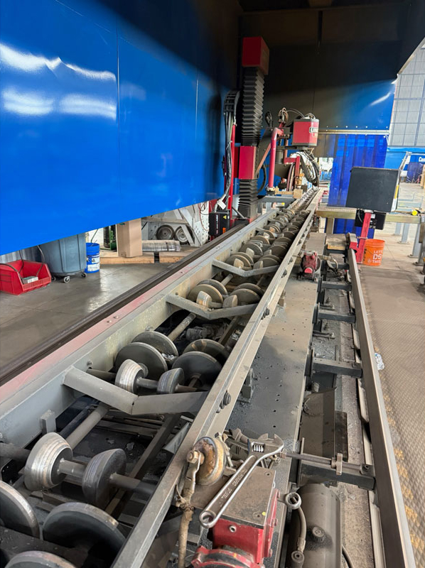 Industrial conveyor with a row of metal rollers under a blue overhead enclosure; factory floor and red maintenance components visible in the background.