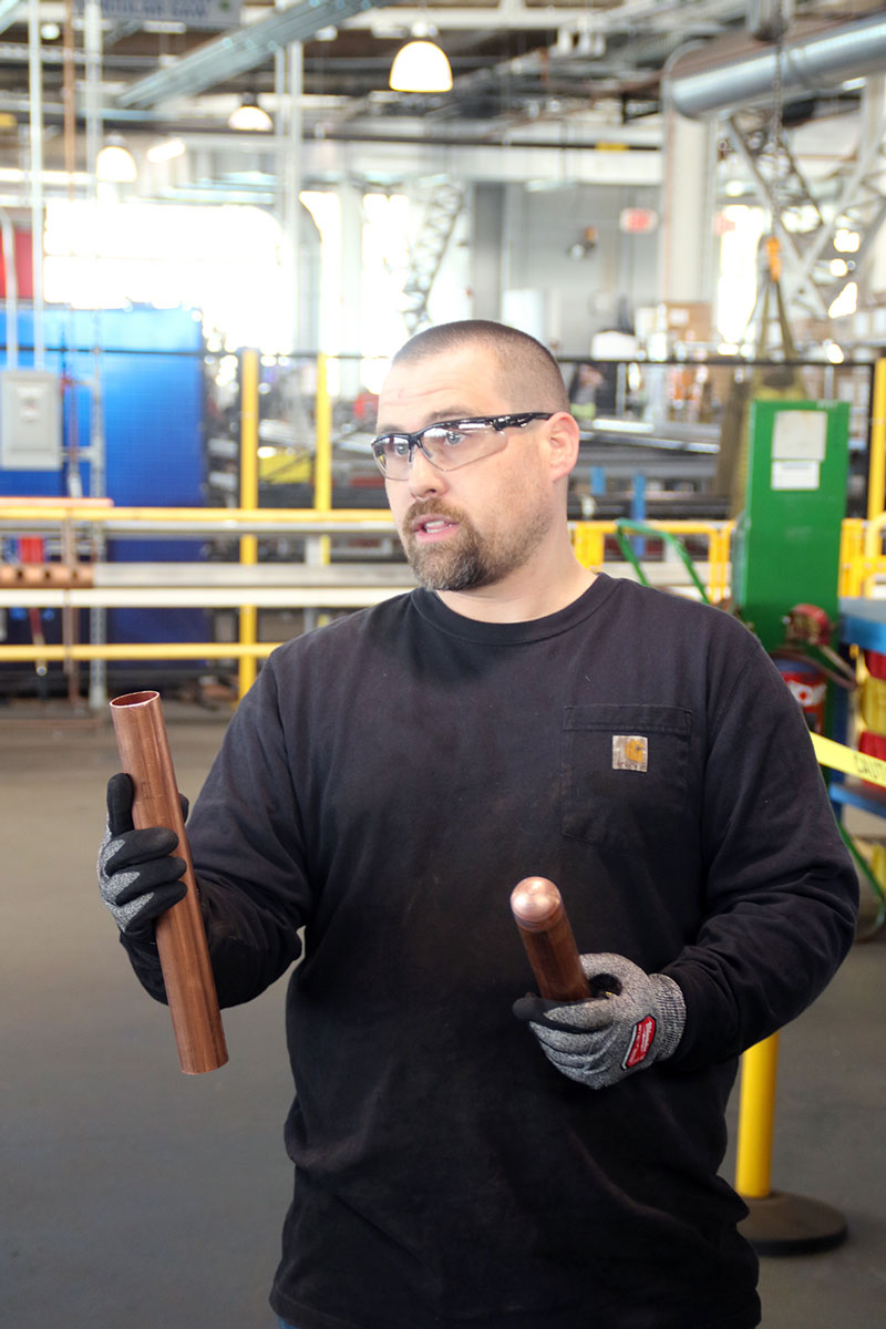 Worker wearing safety glasses and gloves holds copper pipes in an industrial workshop.