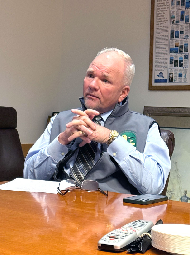 Older man in a vest and tie sits at a conference table, hands clasped, looking to the side with glasses on the desk in front of him.