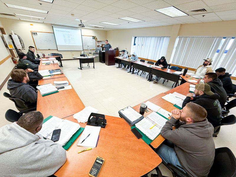 Training session in a classroom: students seated around a U‑shaped set of desks while an instructor presents at the front with a projector screen.