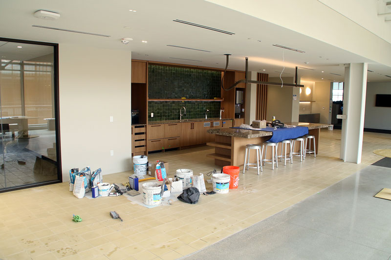 Construction area in a modern kitchenette under renovation; buckets, bags, and tools on the tan tile floor near the island with stools