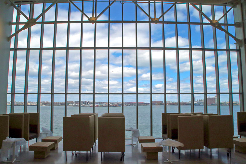 Lounge area with beige chairs and small tables facing a grid of large glass windows overlooking a harbor and city skyline.