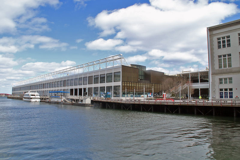 Wide view of a harborfront with a long modern building along the water and a white passenger boat docked at the pier under a blue sky with clouds.