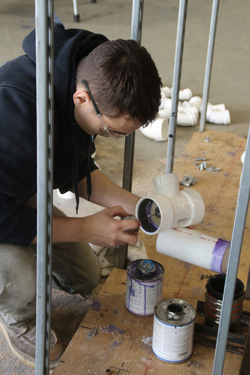 Person kneeling in a workshop, tightening a PVC pipe fitting under a metal frame with paint cans and hardware on a plywood worktable nearby
