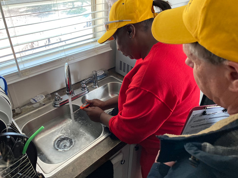 Two volunteers washing dishes at a kitchen sink, wearing yellow caps; one in a red shirt scrubs a utensil while the other watches, clipboard visible nearby with a person photographing or supervising.