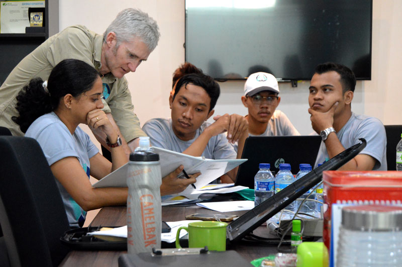 Instructor guides a diverse group of students gathered around a table with papers and laptops in a meeting room.