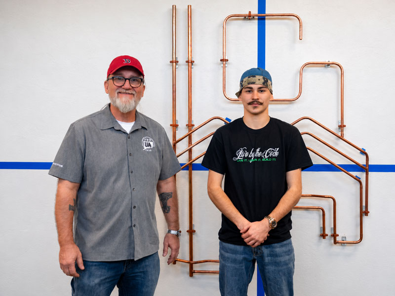 Two men stand side by side in front of a copper pipe art wall installation, posing for a photo in a workshop setting, both smiling at the camera, one wearing a gray shirt and red cap while the other wears a black T-shirt and cap.