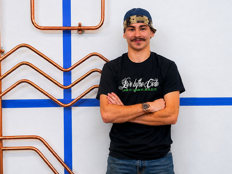 Young man with arms crossed wearing a black t-shirt, standing in front of copper pipes and blue tape lines on a wall.