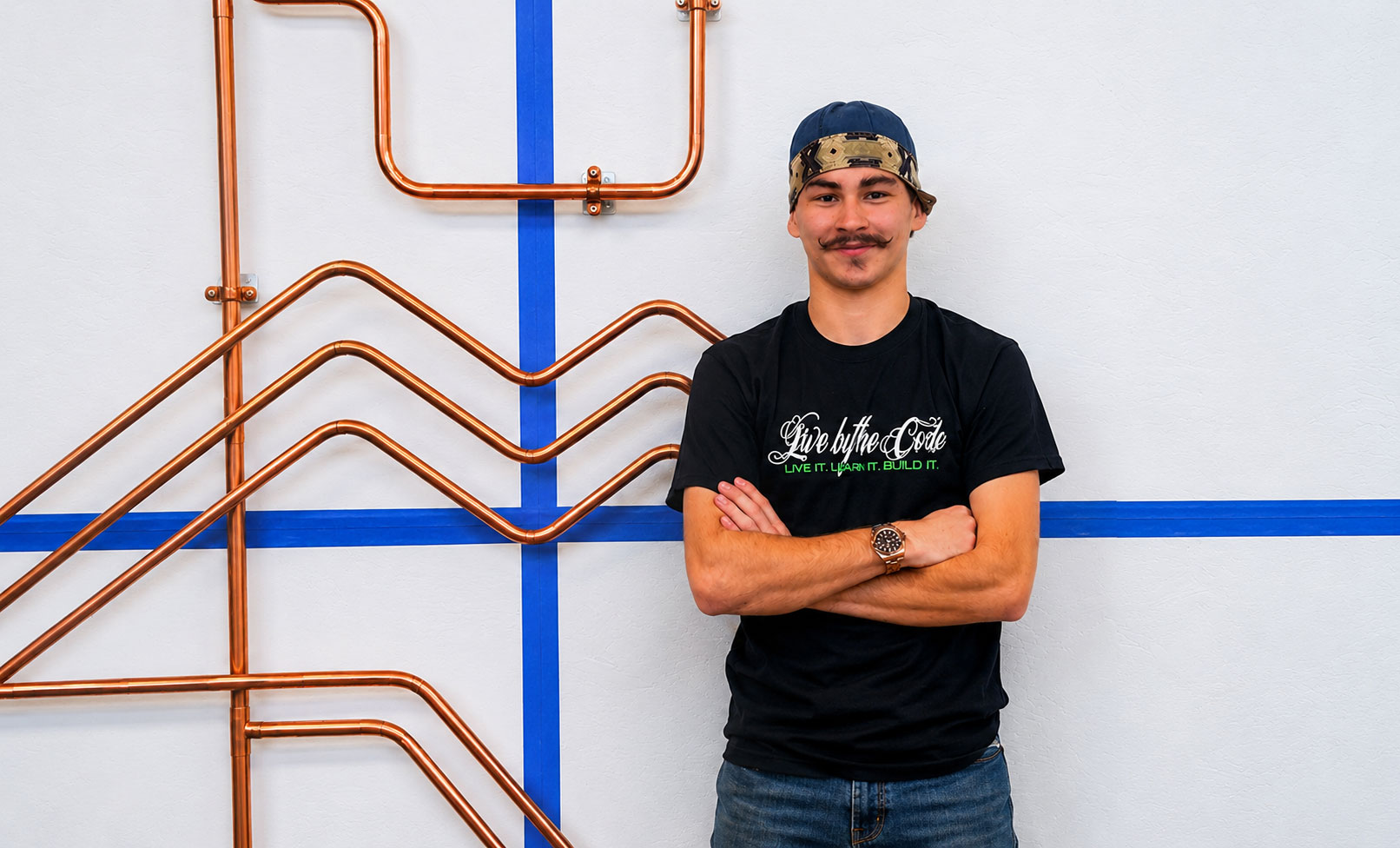 Portrait of a man with a mustache and a backwards cap, arms crossed, standing beside copper pipe artwork on a white wall with blue tape lines. It also shows his black T‑shirt with the slogan 'Live by the Code.'