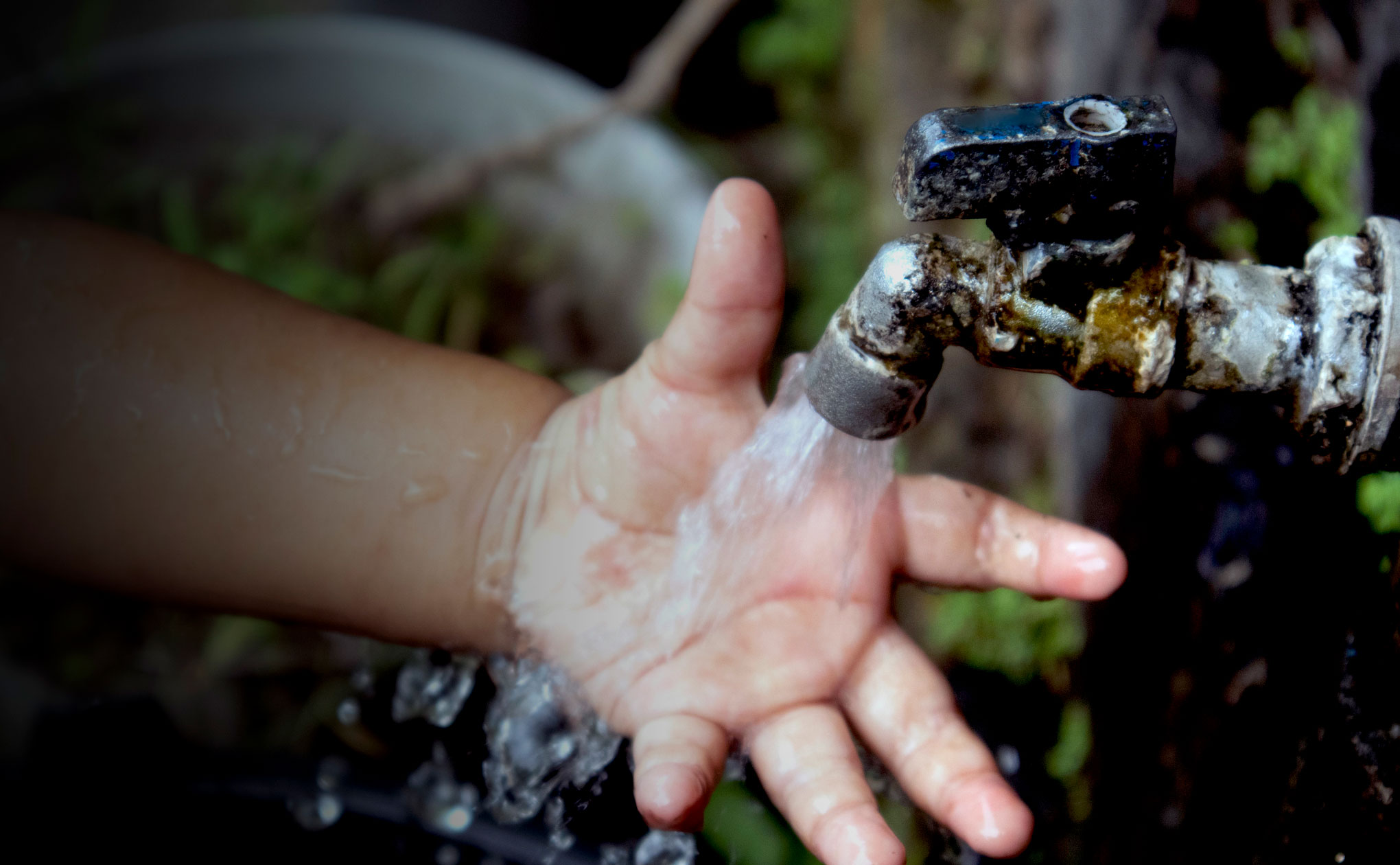 Child's wet hand holds a rusty, corroded pipe valve with water droplets outdoors/near a leak.