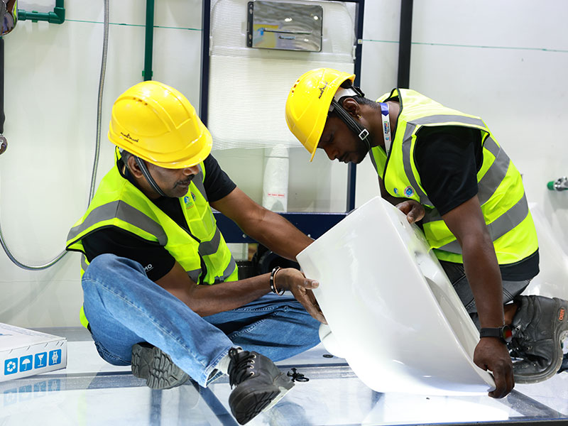 Two construction workers in yellow hard hats and high-visibility vests install a white toilet seat in a building under construction.