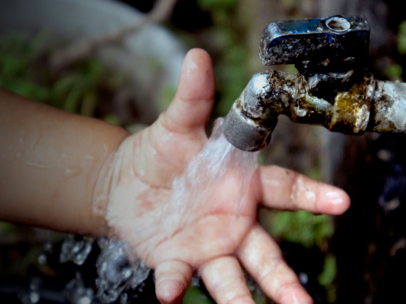 Child's small hand under a running faucet, water pouring over the palm outdoors/nearby greenery.
