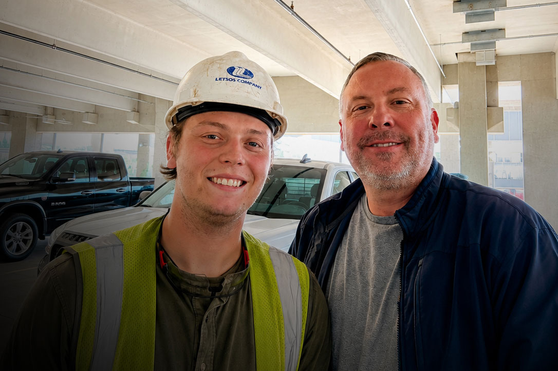 Two men smiling in a construction site parking area, one wearing a hard hat and high‑visibility vest while the other wears a blue jacket behind them on a sunny day in a building under construction.