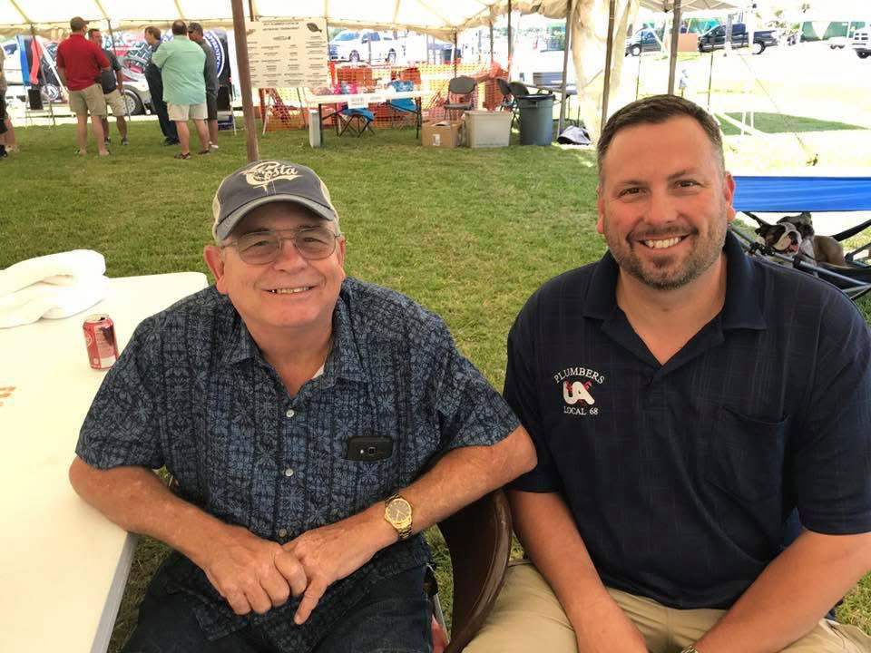 Two men smiling at a table under a white tent at an outdoor event; one wears a cap and glasses, the other a navy polo shirt.