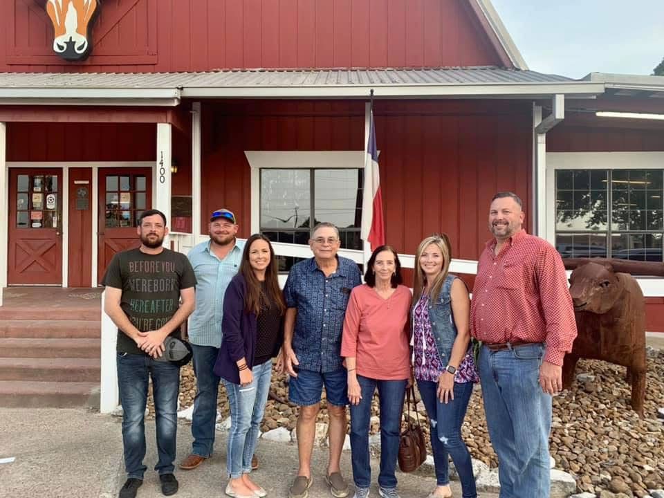 Seven adults pose outside a red barn-style building with a Texas flag and a large animal sculpture nearby.