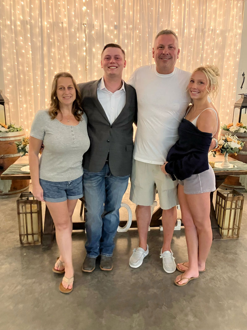 Four adults stand together smiling in front of a twinkling string-light backdrop at a casual gathering.