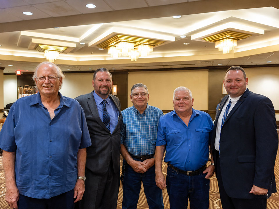 Five men stand side by side in a banquet hall, smiling for the camera; three wear blue shirts and two wear suits.