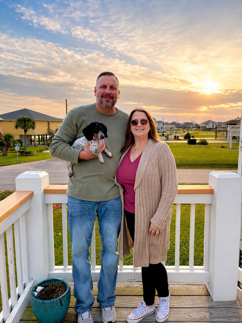 Smiling couple on a deck at sunset, man holding a small black-and-white dog between them.