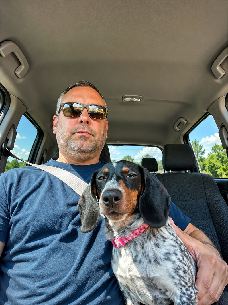 Man wearing sunglasses in a car with a dachshund mix dog on his lap, both facing the camera; seatbelt visible. read more details provided in scene.