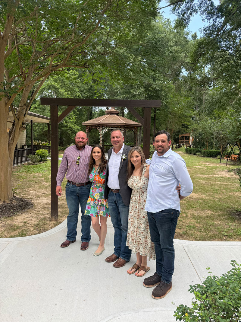 Five adults posing together outdoors at a garden venue, smiling under a wooden arch and gazebo in the background.