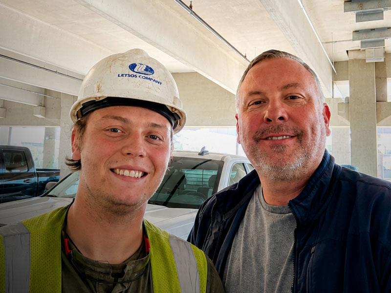 Two men pose for a selfie at a construction site, one wearing a white hard hat and safety vest, the other in a dark jacket.