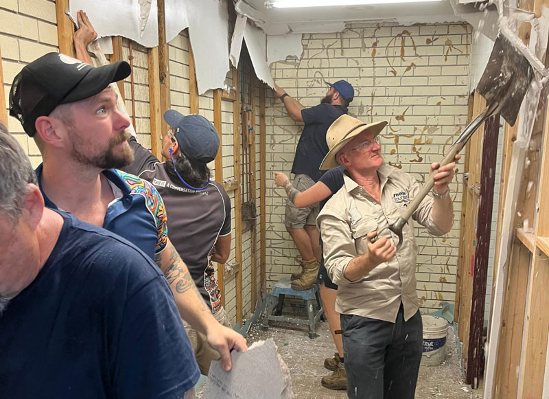 Team of construction workers inside a partially demolished room, removing drywall and tile; man with a hat uses a pry bar.